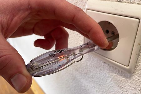 Eine Hand hält einen Spannungsprüfer an eine Steckdose.